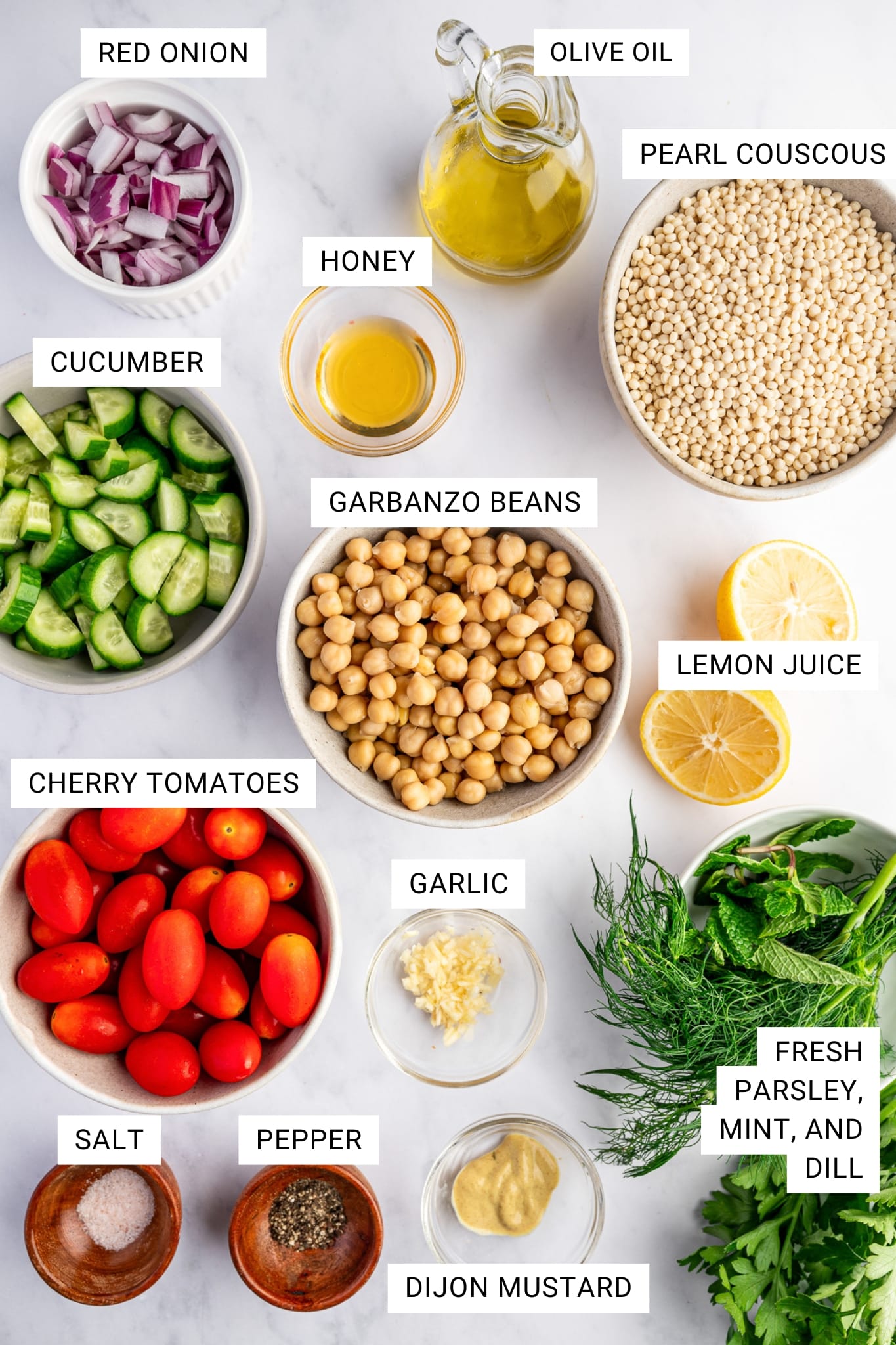 Overhead ingredient shot for pearl couscous salad on a white surface with text overlays labeling each ingredient. Ingredients shown include pearl couscous in a beige bowl labeled “PEARL COUSCOUS,” garbanzo beans in a beige bowl labeled “GARBANZO BEANS,” cucumber in a gray bowl labeled “CUCUMBER,” cherry tomatoes in a beige bowl labeled “CHERRY TOMATOES,” diced red onion in a white ramekin labeled “RED ONION,” olive oil in a glass bottle labeled “OLIVE OIL,” honey in a small glass bowl labeled “HONEY,” lemon halves labeled “LEMON JUICE,” minced garlic in a small glass bowl labeled “GARLIC,” Dijon mustard in a small glass bowl labeled “DIJON MUSTARD,” small wooden bowls labeled “SALT” and “PEPPER,” and fresh parsley, mint, and dill labeled “FRESH PARSLEY, MINT, AND DILL.”