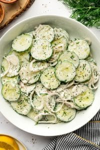 Close overhead view of creamy cucumber salad in a white serving bowl, showing sliced cucumbers and thin shallot rings coated in a creamy white dill dressing and garnished with chopped fresh dill and cracked black pepper, with part of a lemon and striped kitchen towel visible along the edges.
