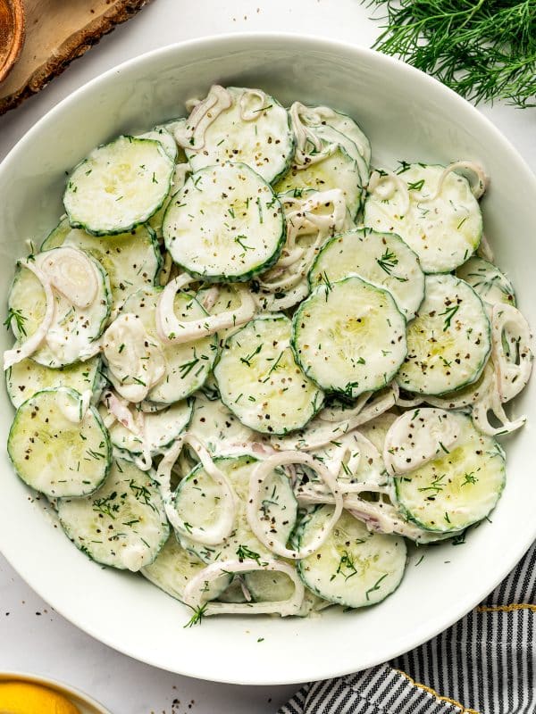 Close overhead view of creamy cucumber salad in a white serving bowl, showing sliced cucumbers and thin shallot rings coated in a creamy white dill dressing and garnished with chopped fresh dill and cracked black pepper, with part of a lemon and striped kitchen towel visible along the edges.