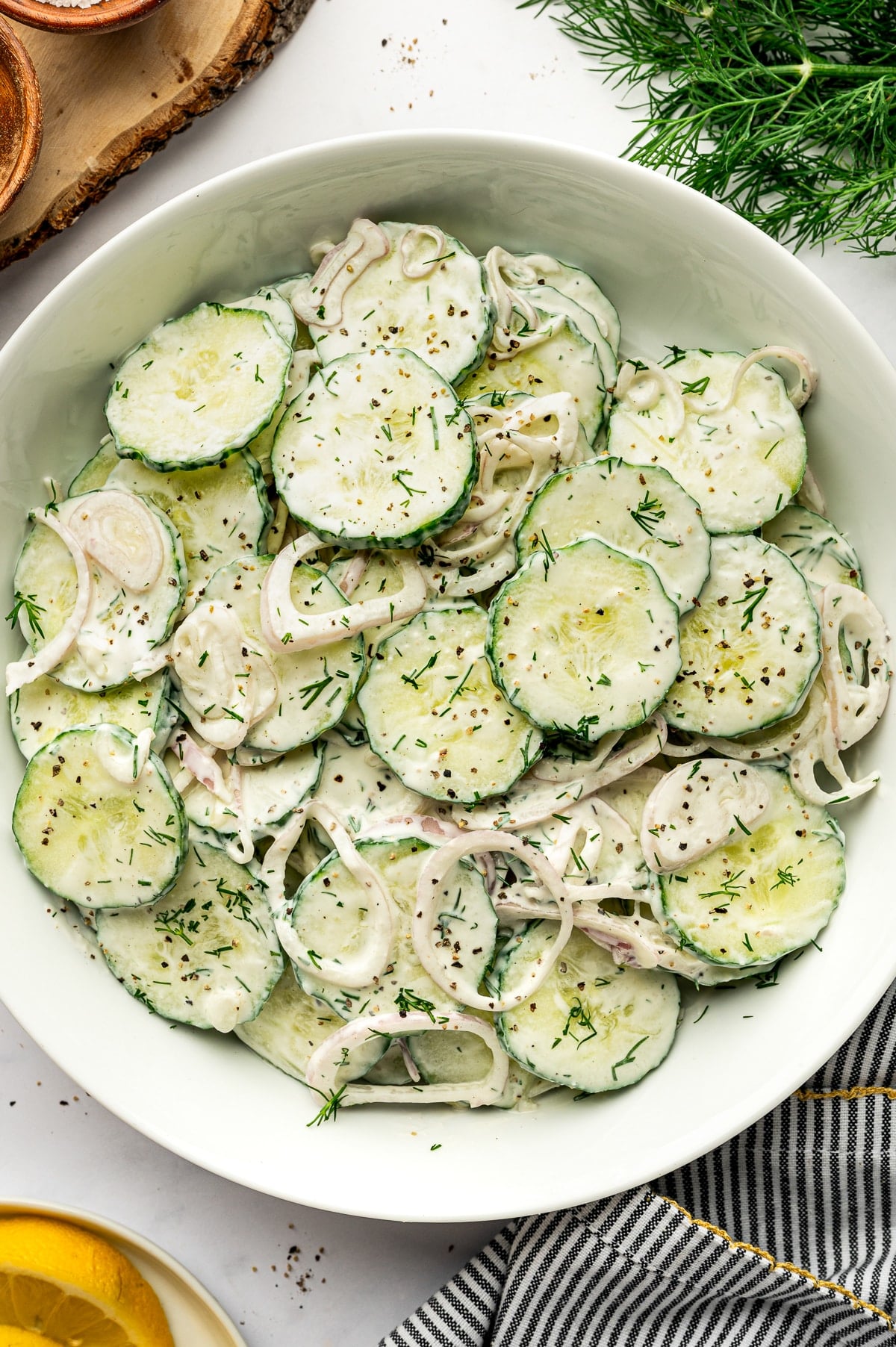 Close overhead view of creamy cucumber salad in a white serving bowl, showing sliced cucumbers and thin shallot rings coated in a creamy white dill dressing and garnished with chopped fresh dill and cracked black pepper, with part of a lemon and striped kitchen towel visible along the edges.