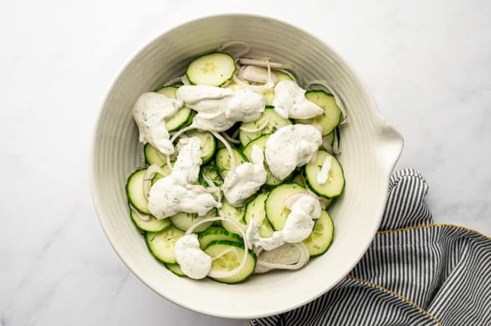 Overhead view of sliced cucumbers and shallots in a large off-white mixing bowl with dollops of creamy dill dressing spooned over the top, set on a light marble background with a black-and-white striped towel trimmed in mustard yellow at the bottom right.