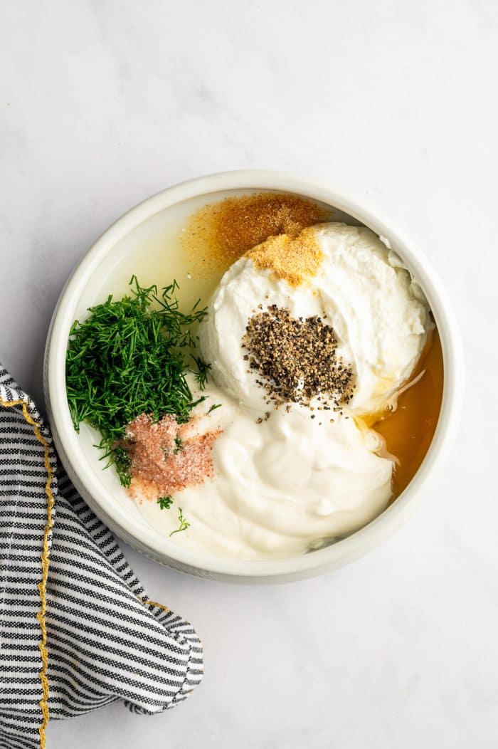 Overhead view of a white bowl filled with the ingredients for creamy cucumber salad dressing, including white Greek yogurt, sour cream, chopped fresh green dill, pale yellow lemon juice, golden honey, black pepper, pink salt, and garlic powder, with a black-and-white striped towel trimmed in mustard yellow beside the bowl on a light marble background.