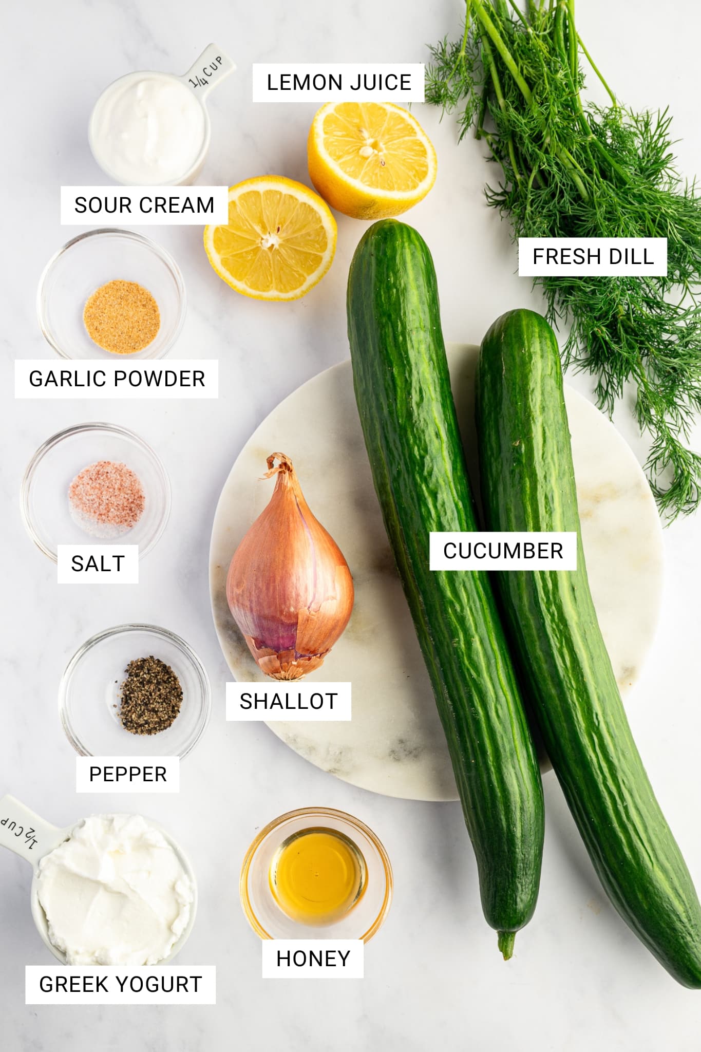 Overhead view of the ingredients for creamy cucumber salad arranged on a light marble background with white text labels naming each ingredient. Two whole dark green cucumbers and one brown shallot sit on a round off-white marble board, surrounded by fresh green dill, two lemon halves, a small glass bowl of golden honey, a white measuring cup of thick white Greek yogurt, a white measuring cup of sour cream, and small glass bowls of garlic powder, pink salt, and black pepper. Visible text overlays read lemon juice, fresh dill, cucumber, shallot, honey, Greek yogurt, pepper, salt, garlic powder, and sour cream.