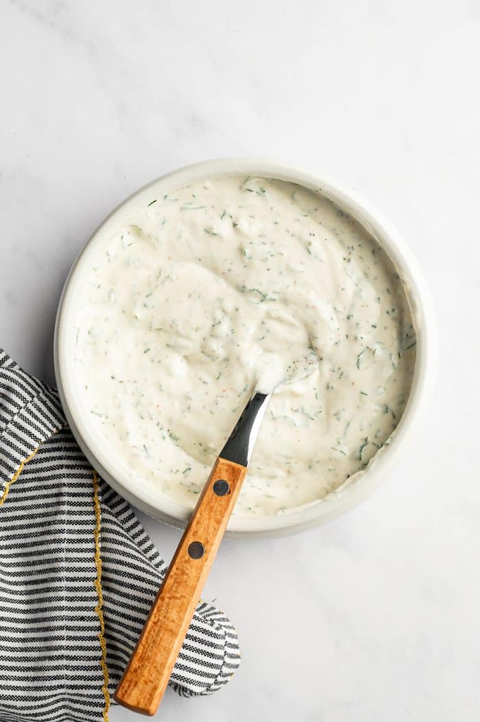 Overhead view of a white bowl filled with smooth creamy dill dressing speckled with green dill and black pepper, with a wooden-handled spoon resting inside the bowl and a black-and-white striped towel with mustard yellow trim on a light marble surface.