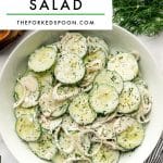 Vertical Pinterest-style graphic for creamy cucumber salad showing a large white bowl filled with sliced cucumbers and thinly sliced shallots coated in a creamy white dill dressing and garnished with chopped fresh dill and cracked black pepper. The bowl sits on a light marble surface with fresh green dill at the top right, part of a black-and-white striped kitchen towel with mustard yellow trim at the bottom right, the edge of a wooden board at the top left, and the rim of a green drinking glass at the bottom left. A large light gray text box at the top reads โCREAMY CUCUMBER SALADโ in bold black letters with โTHEFORKEDSPOON.COMโ underneath and a thin green line between the text.