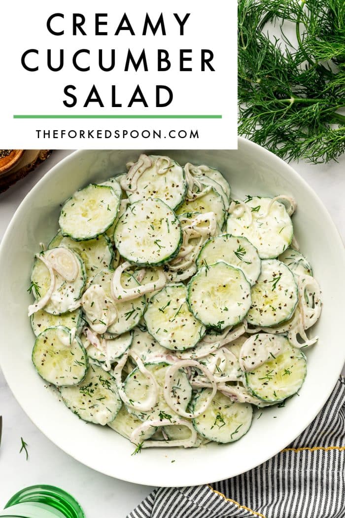 Vertical Pinterest-style graphic for creamy cucumber salad showing a large white bowl filled with sliced cucumbers and thinly sliced shallots coated in a creamy white dill dressing and garnished with chopped fresh dill and cracked black pepper. The bowl sits on a light marble surface with fresh green dill at the top right, part of a black-and-white striped kitchen towel with mustard yellow trim at the bottom right, the edge of a wooden board at the top left, and the rim of a green drinking glass at the bottom left. A large light gray text box at the top reads “CREAMY CUCUMBER SALAD” in bold black letters with “THEFORKEDSPOON.COM” underneath and a thin green line between the text.