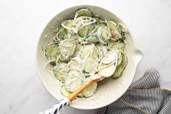 Overhead view of creamy cucumber salad tossed in a large off-white mixing bowl, with sliced cucumbers and shallots coated in a thick white dill dressing and mixed with a white spatula featuring a blue patterned handle, on a light marble surface with a black-and-white striped towel trimmed in mustard yellow.