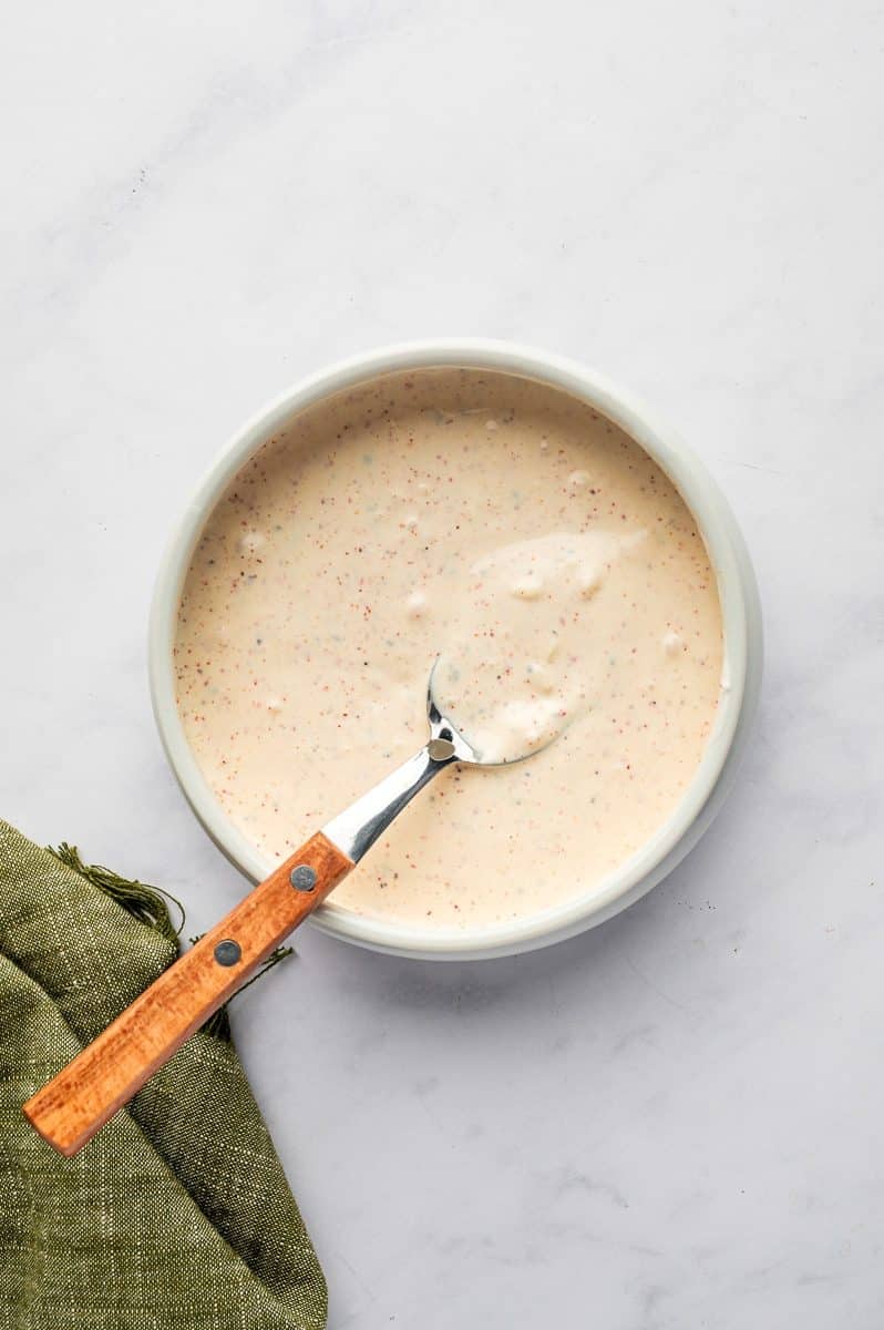 Creamy pale pink lime dressing with red seasoning flecks in a white bowl with a spoon with a wooden handle resting inside, set on a white marble surface with a green linen napkin nearby.