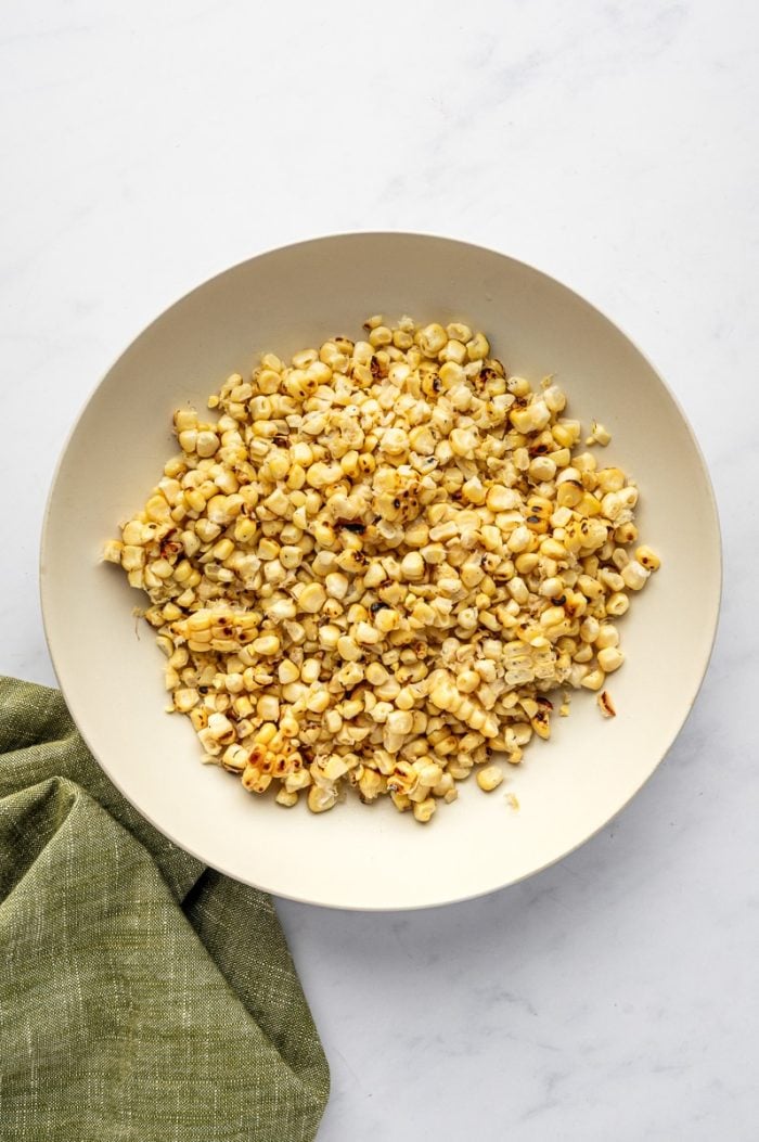 Charred grilled corn kernels in a large cream-colored bowl on a white marble surface with a green linen napkin in the upper left corner.