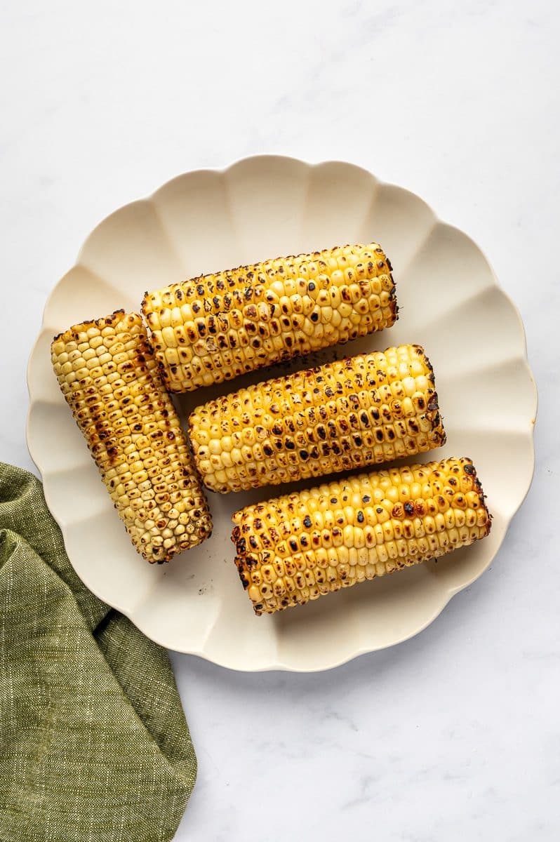 Grilled corn on the cob with charred kernels arranged on a cream-colored scalloped plate on a white marble surface with a green linen napkin in the corner.
