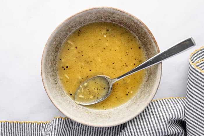 Overhead image of lemon dressing in a speckled beige bowl with a metal spoon resting inside. The dressing is golden yellow with visible black pepper and bits of minced garlic, and the bowl sits on a white surface beside a gray and white striped kitchen towel with yellow trim.