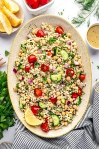 Overhead image of pearl couscous salad on a large oval beige platter with chickpeas, halved cherry tomatoes, cucumber, diced red onion, parsley, mint, and dill. Around the platter are a small white bowl of lemon dressing, lemon wedges in a small bowl, fresh dill, parsley, and a gray and white striped towel with yellow trim on a white background.