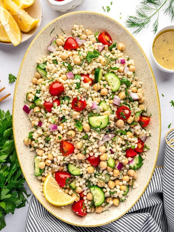 Overhead image of pearl couscous salad on a large oval beige platter with chickpeas, halved cherry tomatoes, cucumber, diced red onion, parsley, mint, and dill. Around the platter are a small white bowl of lemon dressing, lemon wedges in a small bowl, fresh dill, parsley, and a gray and white striped towel with yellow trim on a white background.
