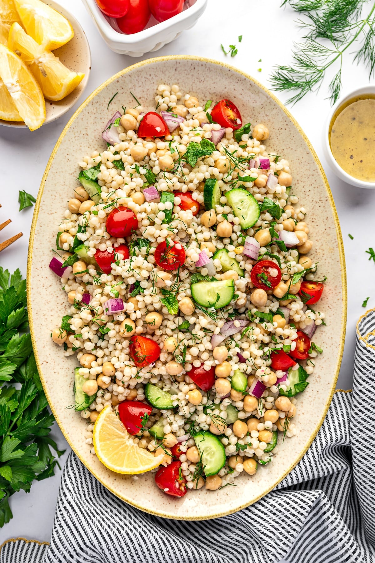 Overhead image of pearl couscous salad on a large oval beige platter with chickpeas, halved cherry tomatoes, cucumber, diced red onion, parsley, mint, and dill. Around the platter are a small white bowl of lemon dressing, lemon wedges in a small bowl, fresh dill, parsley, and a gray and white striped towel with yellow trim on a white background.