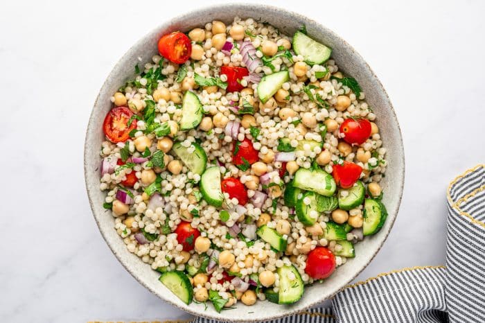 Overhead image of mixed pearl couscous salad in a light beige bowl with chickpeas, halved cherry tomatoes, chopped cucumber, diced red onion, parsley, mint, and dill evenly tossed together. The bowl sits on a white surface next to a gray and white striped kitchen towel with yellow trim.