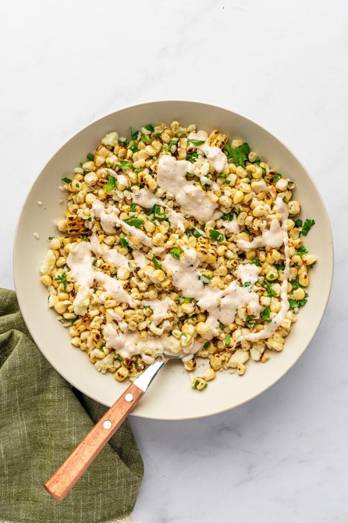Grilled corn kernels, crumbled cotija cheese, chopped cilantro, diced jalapeño, and creamy dressing in a large cream-colored bowl with a spoon with a wooden handle on a white marble surface and a green linen napkin nearby.
