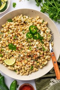 Mexican street corn salad in a large cream-colored bowl topped with crumbled cotija cheese, chopped cilantro, red chile seasoning, sliced jalapeños, and a lime wedge, with fresh cilantro, lime wedges, whole jalapeños, a small wooden bowl of red seasoning, a spoon with a wooden handle, and a green linen napkin around the bowl.