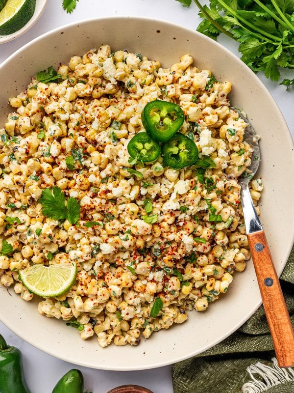 Mexican street corn salad in a large cream-colored bowl topped with crumbled cotija cheese, chopped cilantro, red chile seasoning, sliced jalapeños, and a lime wedge, with fresh cilantro, lime wedges, whole jalapeños, a small wooden bowl of red seasoning, a spoon with a wooden handle, and a green linen napkin around the bowl.