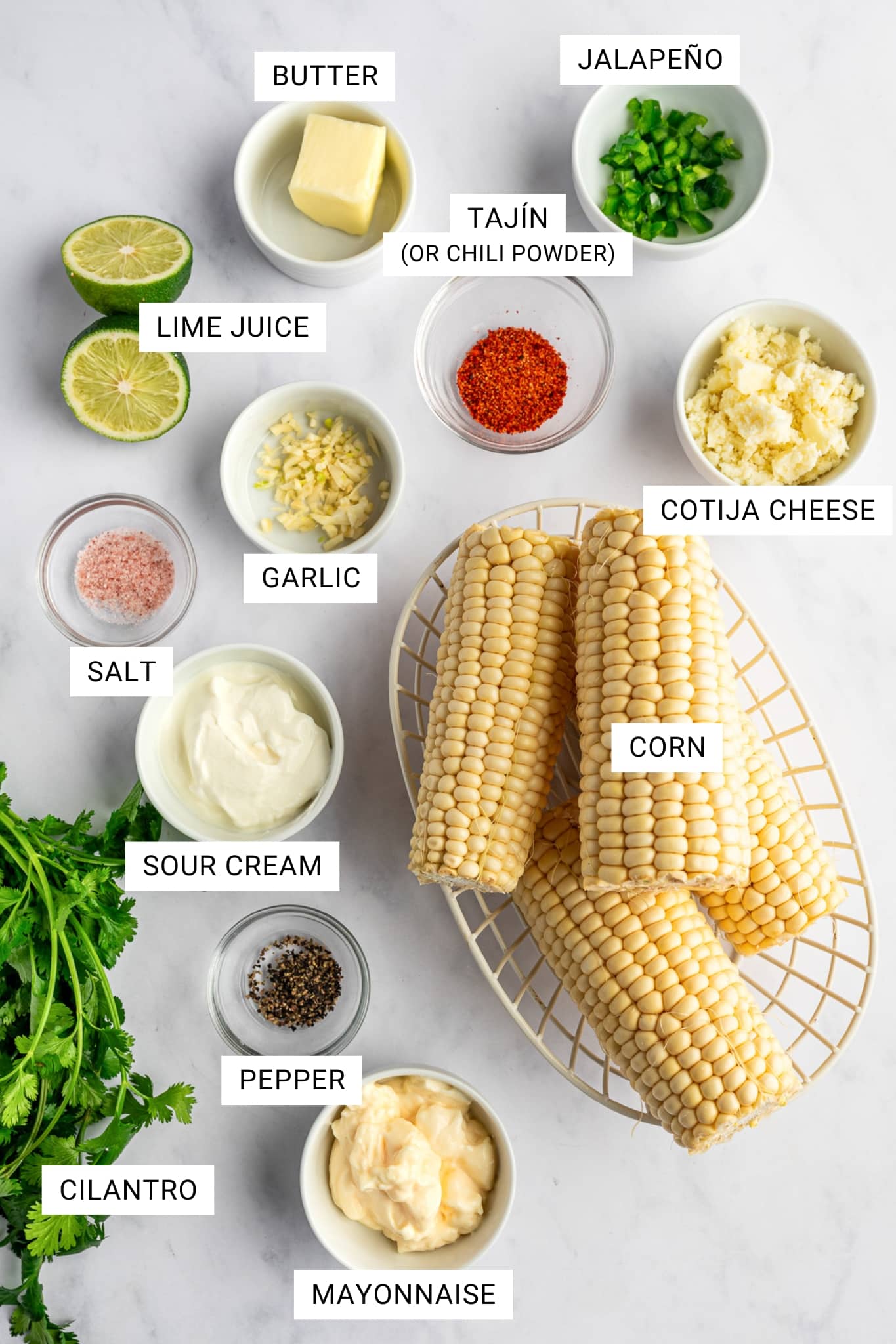 Ingredients for Mexican street corn salad arranged on a white marble surface, including corn on the cob in a cream wire basket, cilantro, mayonnaise, sour cream, salt, pepper, garlic, lime juice, butter, Tajín or chili powder, jalapeño, and cotija cheese in small bowls, with white text labels identifying each ingredient.