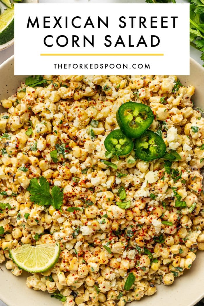 Pinterest pin image of Mexican street corn salad in a cream-colored bowl topped with sliced jalapeños, crumbled cotija cheese, chopped cilantro, red seasoning, and a lime wedge, with black text on a white banner reading “MEXICAN STREET CORN SALAD” and “THEFORKEDSPOON.COM.”