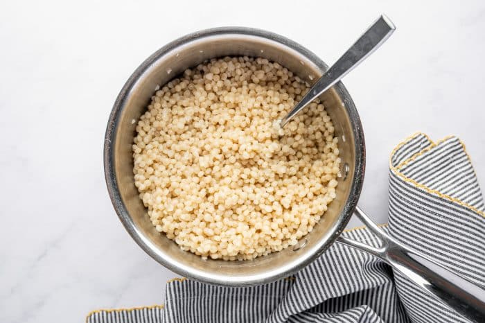 Overhead image of cooked pearl couscous in a silver saucepan with a metal spoon resting inside. The saucepan sits on a white surface next to a gray and white striped kitchen towel with yellow trim.