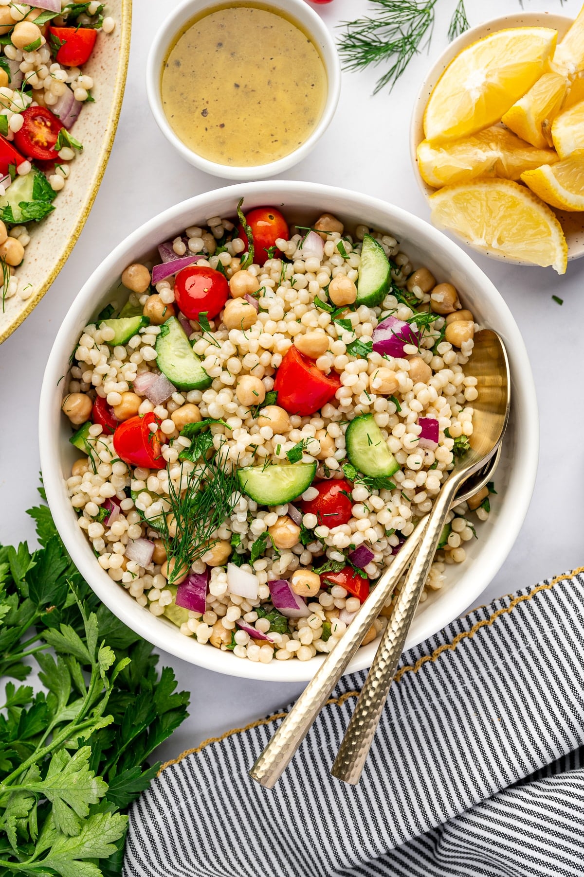 Overhead image of pearl couscous salad in a round white bowl with gold serving spoons tucked inside. The salad includes chickpeas, cherry tomatoes, cucumber, diced red onion, parsley, mint, and dill. In the background are a small white bowl of dressing, lemon wedges, part of a serving platter, fresh parsley, and a gray and white striped towel with yellow trim.