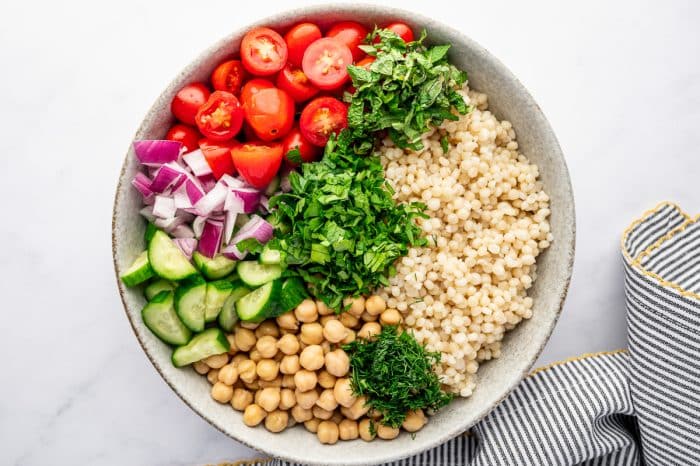 Overhead image of a large light beige bowl filled with pearl couscous, chickpeas, halved cherry tomatoes, chopped cucumber, diced red onion, chopped parsley, chopped mint, and fresh dill arranged in sections before mixing. The bowl sits on a white surface next to a gray and white striped kitchen towel with yellow trim.