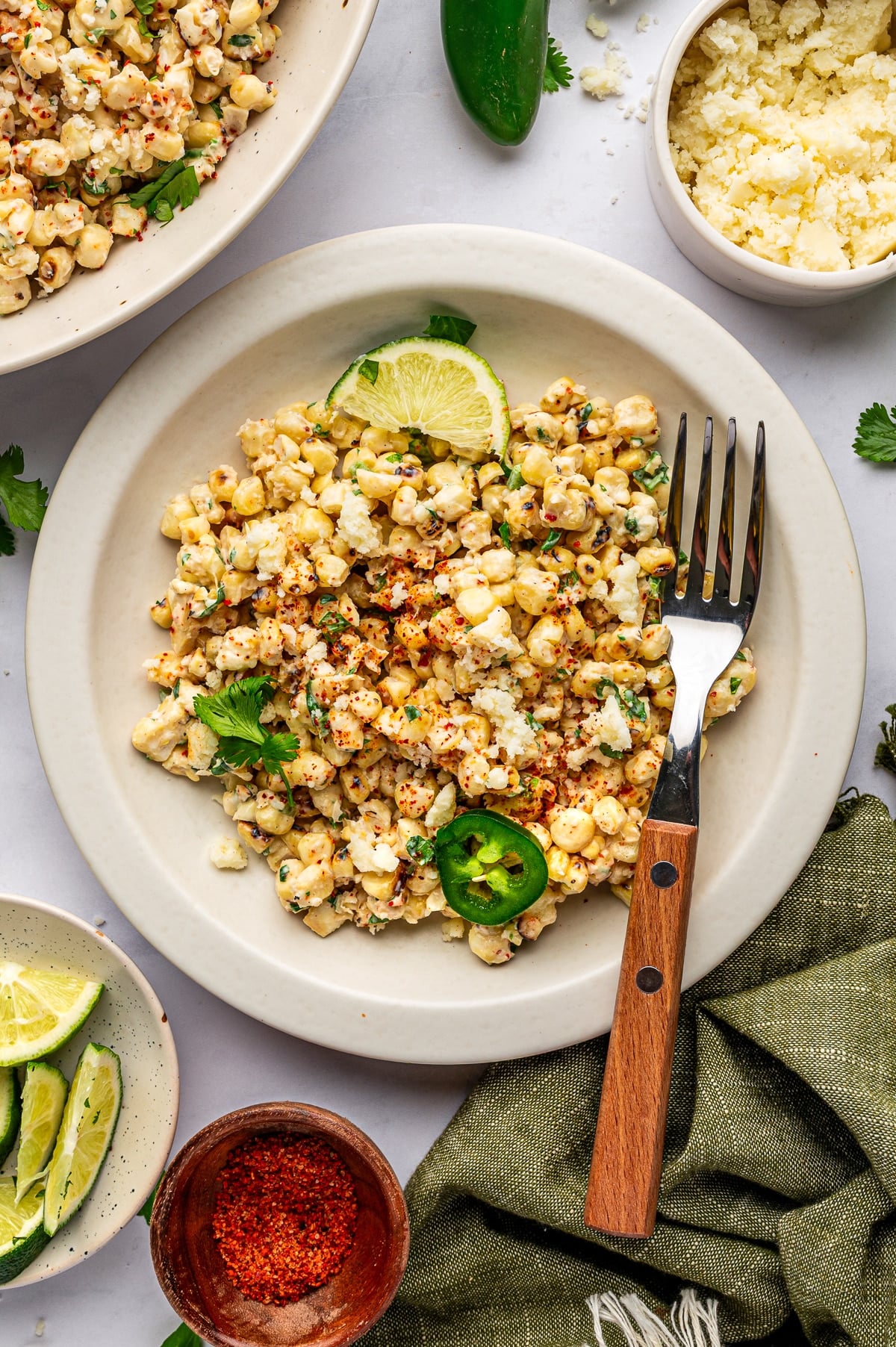 A serving of Mexican street corn salad on a cream-colored plate topped with crumbled cotija cheese, chopped cilantro, red chile seasoning, a lime wedge, and sliced jalapeño, with a fork with a wooden handle, lime wedges, fresh cilantro, whole jalapeños, a white bowl of cotija cheese, a small wooden bowl of red seasoning, a large bowl of corn salad, and a green linen napkin nearby.