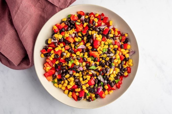 Overhead image of black bean and corn salad ingredients mixed together in a large beige bowl, with black beans, yellow corn, diced red bell pepper, chopped red onion, and green jalapeño on a light gray surface next to a mauve linen napkin.