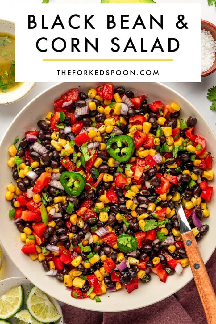 Pinterest pin image for black bean and corn salad showing a large beige bowl filled with black beans, yellow corn, diced red bell pepper, red onion, jalapeño, and cilantro with a wooden-handled serving spoon. A white text box overlay at the top reads “BLACK BEAN & CORN SALAD” with “THEFORKEDSPOON.COM” underneath and a thin yellow line between the title and website.
