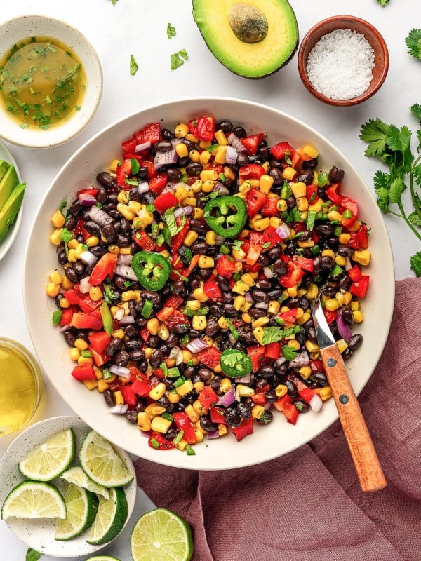 Overhead image of black bean and corn salad in a large beige serving bowl topped with sliced jalapeño and cilantro, surrounded by lime wedges, a halved avocado, coarse salt in a small brown bowl, cumin lime dressing, olive oil, sliced avocado, fresh cilantro, and a mauve linen napkin.