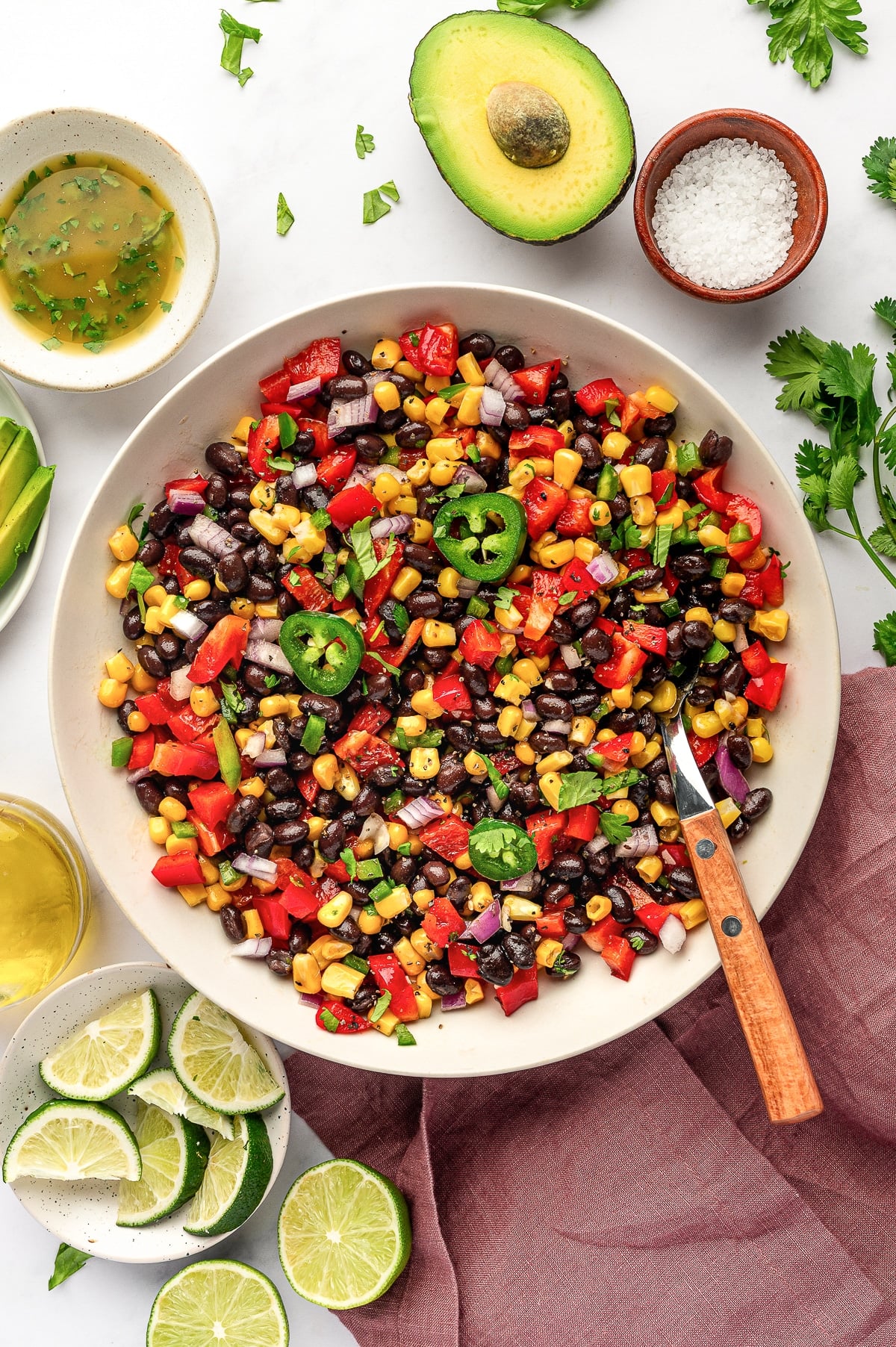 Overhead image of black bean and corn salad in a large beige serving bowl topped with sliced jalapeño and cilantro, surrounded by lime wedges, a halved avocado, coarse salt in a small brown bowl, cumin lime dressing, olive oil, sliced avocado, fresh cilantro, and a mauve linen napkin.