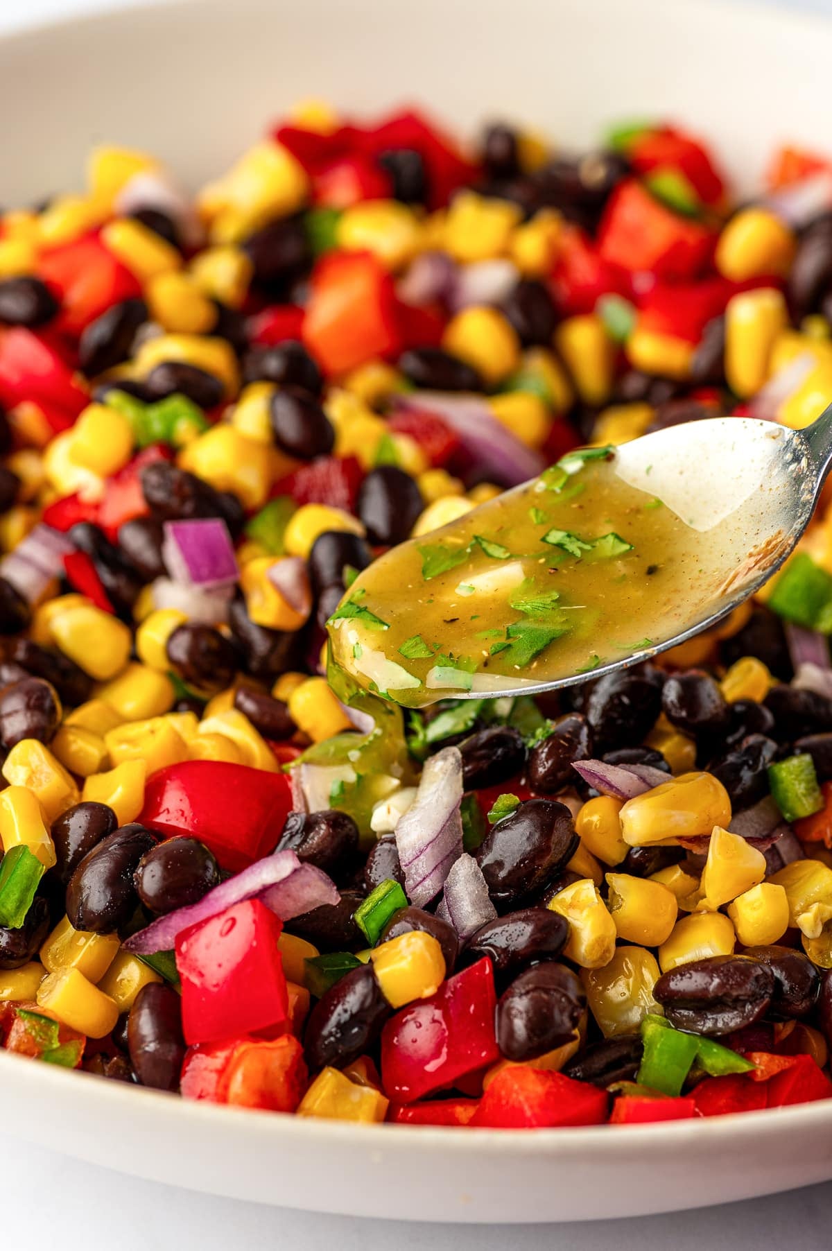 Close-up image of a spoonful of cumin lime dressing being poured over black bean and corn salad in a beige bowl, with black beans, yellow corn, diced red bell pepper, red onion, jalapeño, and cilantro visible in the background.