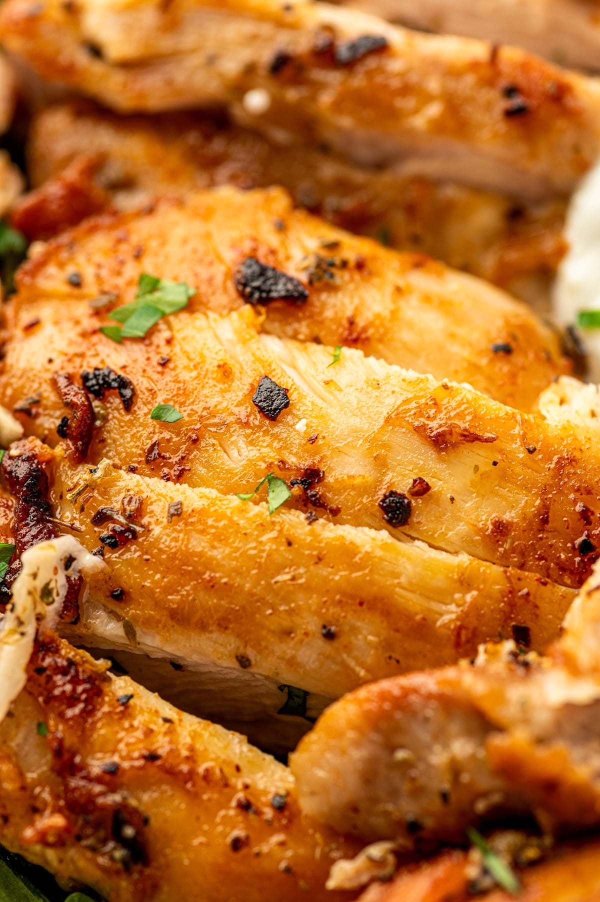 Extreme close-up of sliced Greek chicken thighs with golden browned edges, visible garlic, black pepper, dried herbs, and small pieces of fresh parsley.