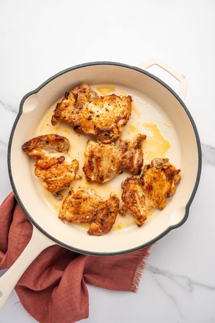 Overhead view of golden brown Greek chicken thighs cooked in a cream-colored skillet with lightly charred edges and pan juices, set on a white marble surface with a rust-colored cloth napkin underneath.