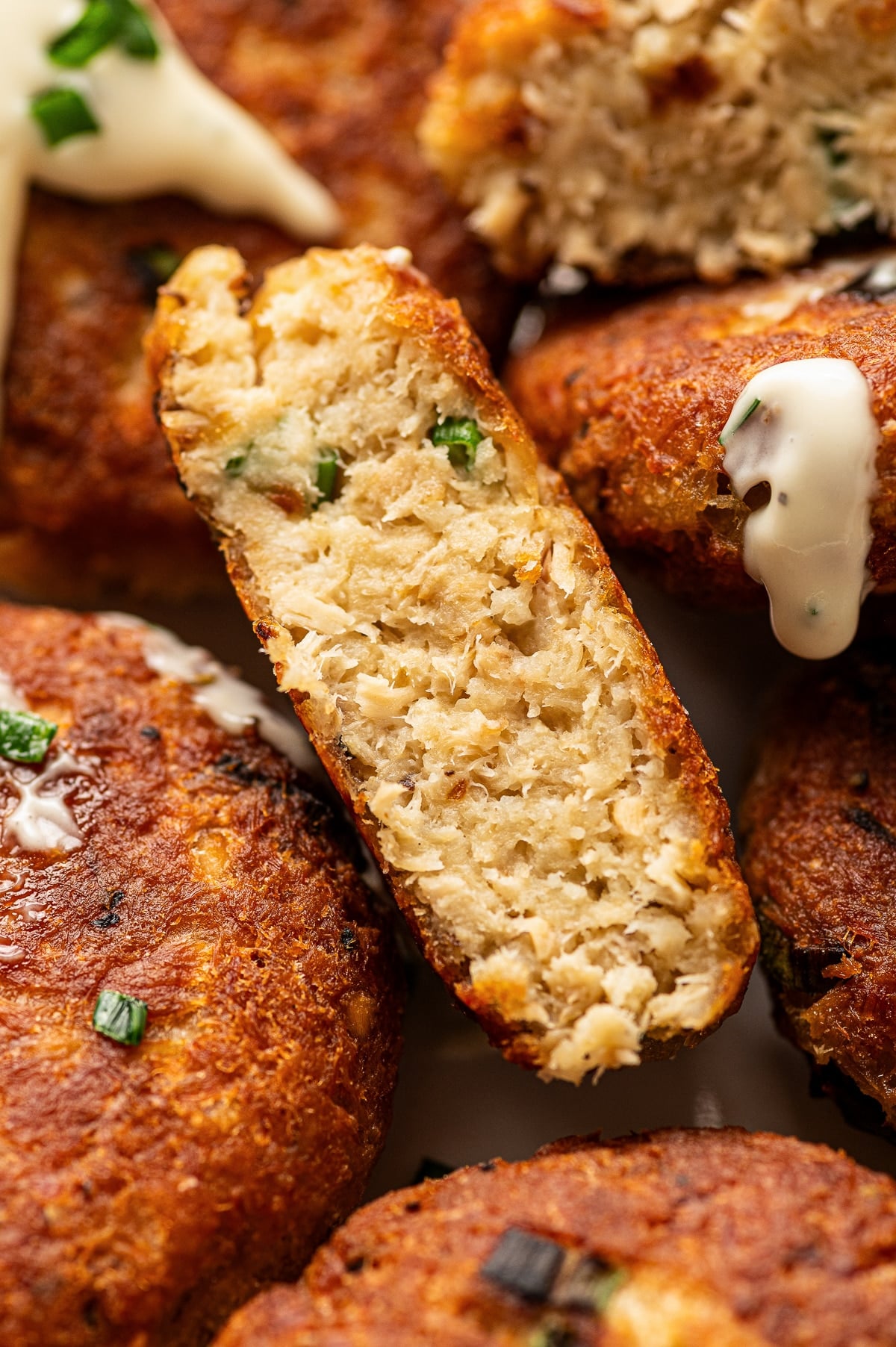 Close-up of crispy golden brown salmon cakes with one salmon cake cut in half to show the tender, flaky salmon filling inside. The cakes are topped with creamy white sauce and small pieces of chopped green chives, with browned edges and visible bits of green onion.