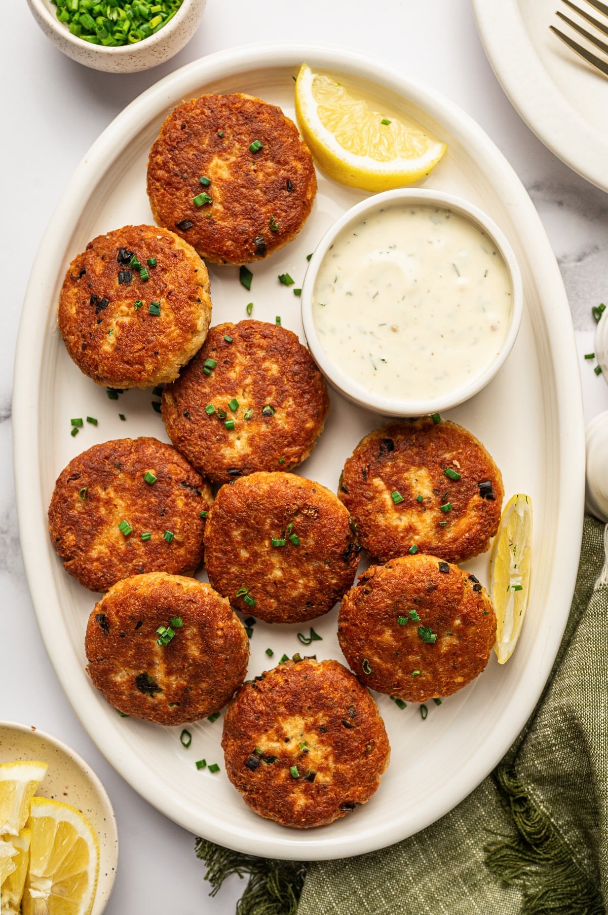 Crispy golden salmon cakes arranged on a cream-colored oval platter with a small white bowl of creamy tartar sauce, lemon wedges, chopped green chives, an olive-green napkin, and a small bowl of lemon wedges on a white marble surface.