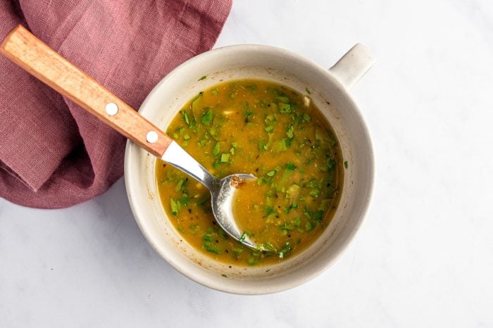 Overhead image of cumin lime dressing in a small beige bowl with chopped cilantro, garlic, lime juice, olive oil, cumin, salt, and black pepper mixed together, with a metal spoon with a wooden handle resting inside and a mauve linen napkin on the side.