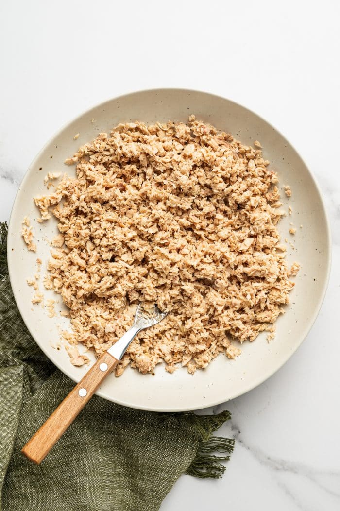 Flaked canned salmon in a shallow, beige-speckled bowl with a wooden-handled fork, set on a white marble surface with an olive-green linen napkin.