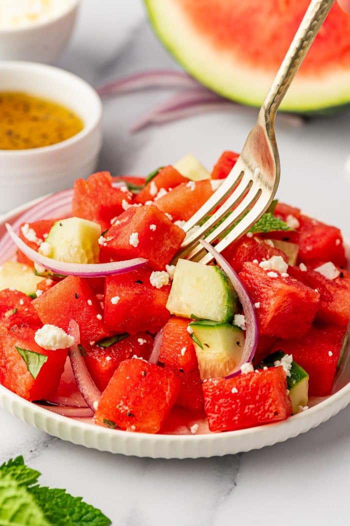 A silver fork lifting a bite of watermelon cucumber salad from a small white bowl filled with red watermelon cubes, chopped cucumber, sliced red onion, crumbled feta cheese, fresh herbs, and black pepper, with honey-lime dressing and a watermelon wedge in the background.