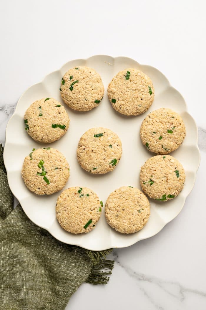 Nine uncooked salmon cake patties are arranged on a cream-colored scalloped plate, with small pieces of green onion visible throughout the pale salmon mixture, and set on a white marble surface beside an olive-green napkin.