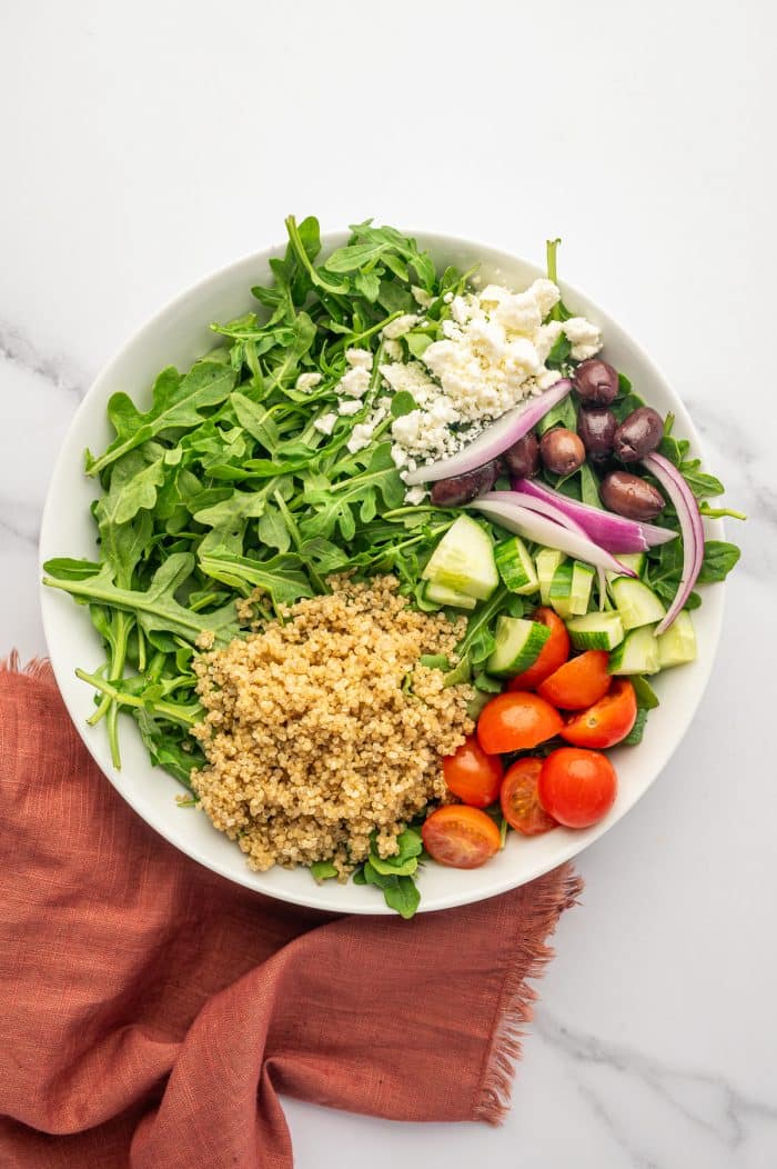 Overhead view of Greek chicken bowl ingredients arranged in a white bowl, including cooked quinoa, fresh arugula, diced cucumber, halved cherry tomatoes, sliced red onion, Kalamata olives, and crumbled feta cheese on a white marble surface with a rust-colored cloth napkin.