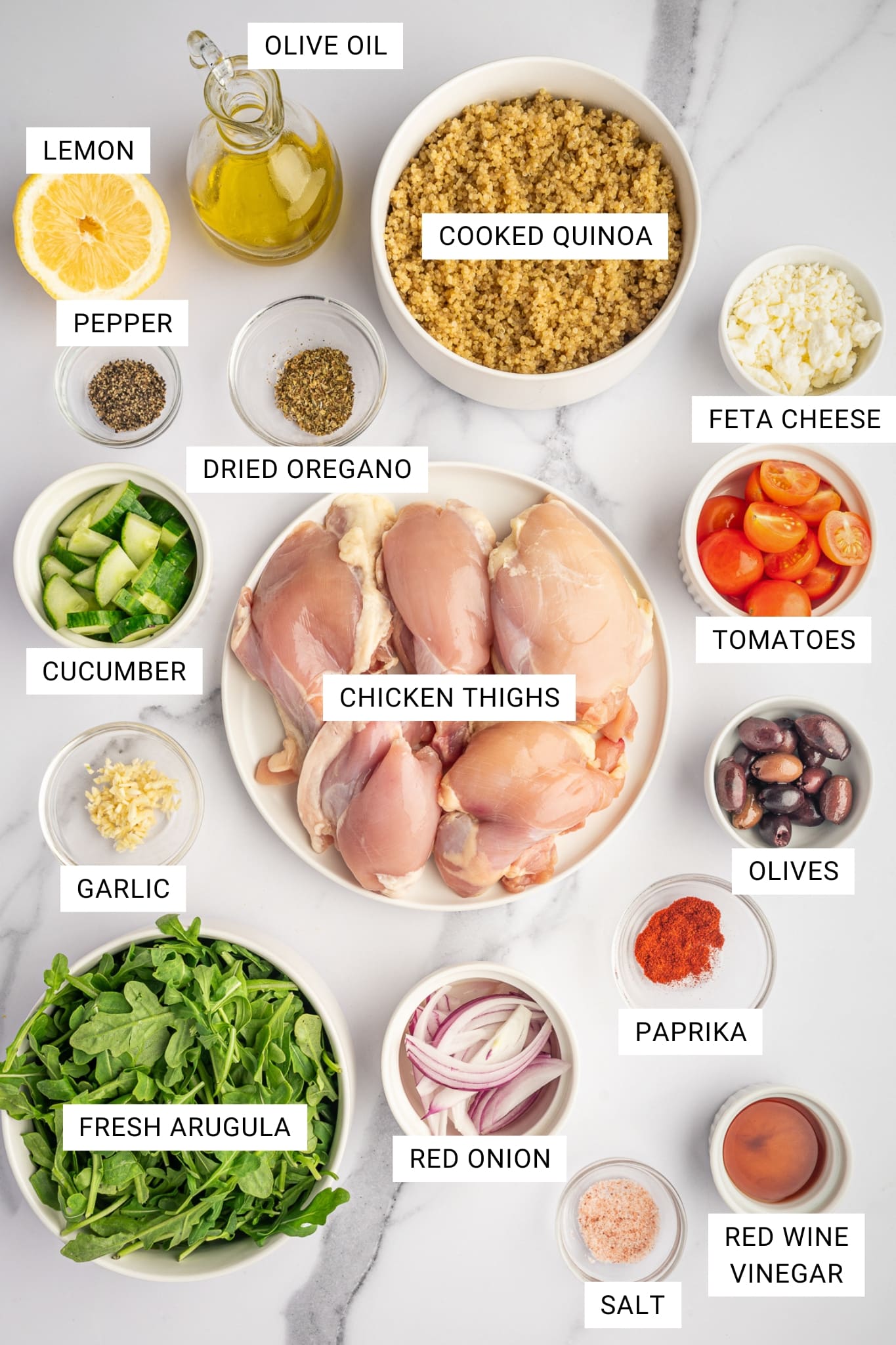 Overhead labeled ingredient photo for Greek chicken bowls on a white marble surface with text overlays for lemon, olive oil, cooked quinoa, feta cheese, pepper, dried oregano, tomatoes, cucumber, chicken thighs, olives, garlic, fresh arugula, red onion, paprika, red wine vinegar, and salt.