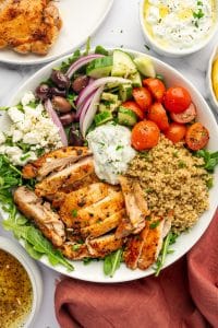 Overhead view of a Greek chicken bowl in a white bowl with sliced golden chicken, cooked quinoa, arugula, cherry tomatoes, diced cucumber, red onion, Kalamata olives, crumbled feta cheese, and a spoonful of tzatziki, with extra dressing, chicken, and a rust-colored cloth napkin nearby.