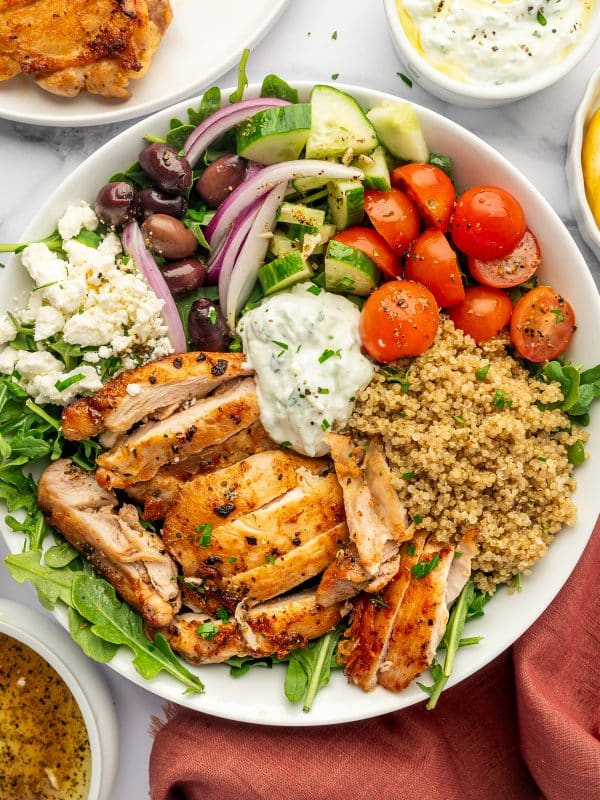 Overhead view of a Greek chicken bowl in a white bowl with sliced golden chicken, cooked quinoa, arugula, cherry tomatoes, diced cucumber, red onion, Kalamata olives, crumbled feta cheese, and a spoonful of tzatziki, with extra dressing, chicken, and a rust-colored cloth napkin nearby.