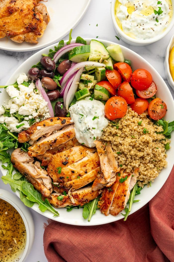 Overhead view of a Greek chicken bowl in a white bowl with sliced golden chicken, cooked quinoa, arugula, cherry tomatoes, diced cucumber, red onion, Kalamata olives, crumbled feta cheese, and a spoonful of tzatziki, with extra dressing, chicken, and a rust-colored cloth napkin nearby.