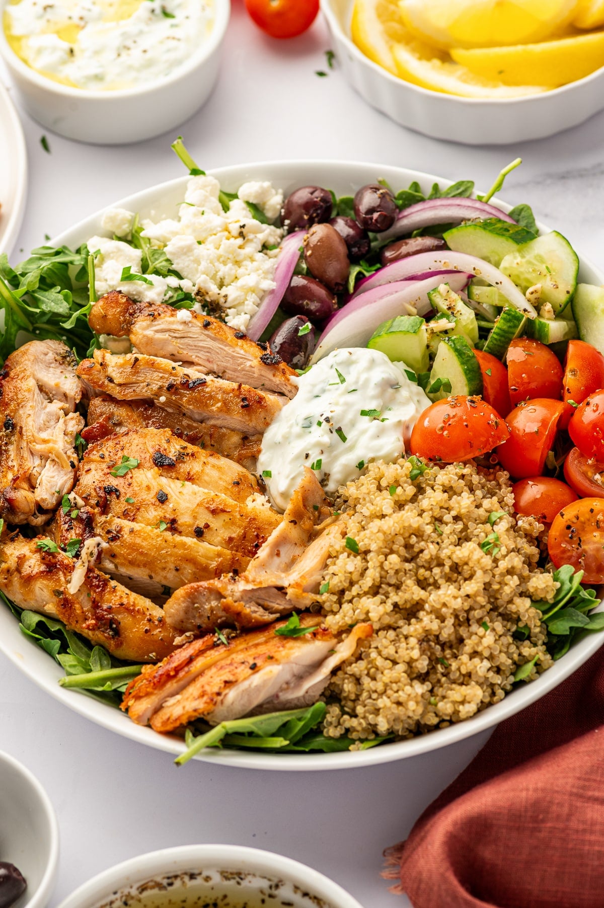 Close-up of a Greek chicken quinoa bowl with sliced golden chicken, cooked quinoa, cherry tomatoes, diced cucumber, red onion, Kalamata olives, crumbled feta cheese, arugula, and a spoonful of tzatziki, with lemon wedges and extra sauce in the background.