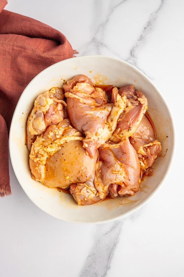 Overhead view of raw boneless chicken thighs coated in a lemon-oregano marinade inside a white bowl on a white marble surface, with a rust-colored cloth napkin on the left side.