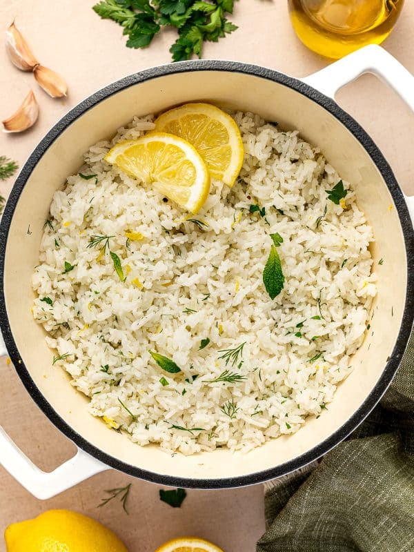 Fluffy Greek lemon rice with chopped parsley, dill, mint, and lemon zest in a cream-colored Dutch oven, garnished with lemon slices. Fresh parsley, garlic cloves, olive oil, whole lemons, lemon halves, and a green napkin are arranged around the pot on a beige surface.