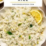 Pinterest pin image showing Greek lemon rice in a white scalloped bowl with lemon slices, fresh herbs, lemon zest, and black pepper. A white text box overlay reads โGREEK LEMON RICEโ with โTHEFORKEDSPOON.COMโ beneath it and a pale yellow underline.